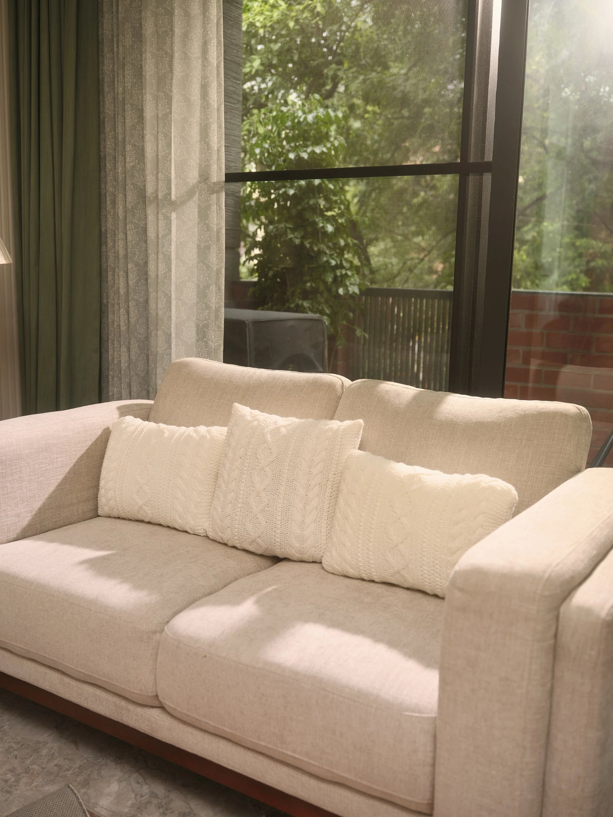 The Maman Cable-Knit Cushion Covers by Uahan — three ivory cable-knit cushions on a linen sofa by the window